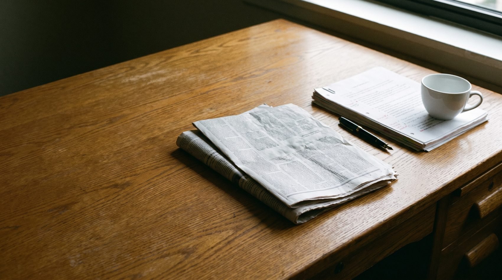 A newspaper, fountain pen and marked-up research memo on an oak desk
